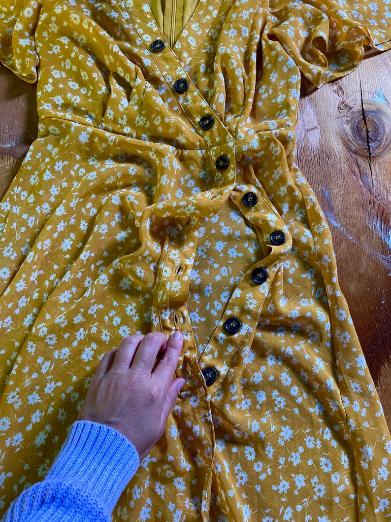 May include: A yellow floral wrap dress with white flowers and a row of buttons down the front.
