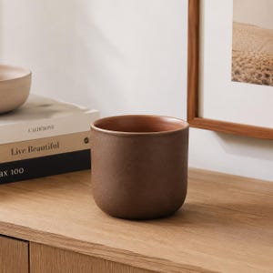 May include: A cylindrical ceramic planter with a matte, dark brown exterior finish and a smooth, lighter terracotta-coloured interior. The vessel features a simple, straight-sided design with a rounded base, presented as a standalone decorative object.
