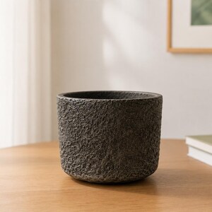 May include: A cylindrical planter with a dark grey, heavily textured, rough exterior surface. The pot features a wide, open top and a flat base, showcasing a rugged, stone-like appearance with deep crevices and an uneven, tactile finish.