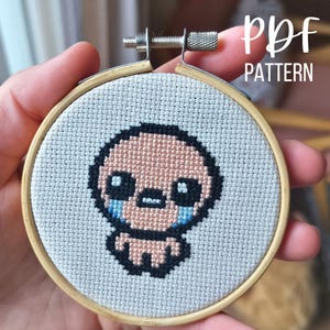 Puede incluir: Un bordado de punto de cruz en un aro de madera presenta un personaje pixelado con una cara rosa pálido, ojos negros y lágrimas azules. El texto "PDF PATTERN" está en escritura blanca. El bordado se sostiene en una mano.