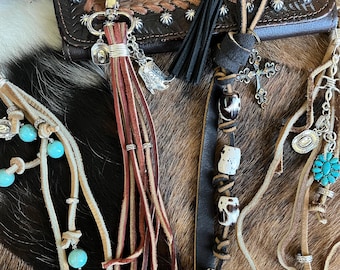 Op de afbeelding: Een collectie leren sleutelhangers met lange franjes, turkoois kralen, zilverkleurige bedels waaronder een cowboyhoed en laars, een zwarte kwast, een kruishanger en witte kralen met zwarte vlekken, bevestigd aan metalen ringen en clips.