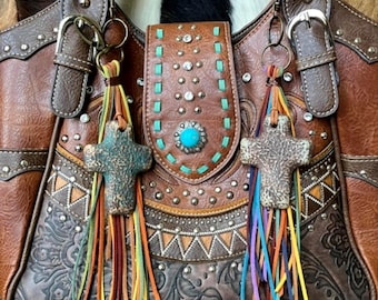 Op de afbeelding: Een bruine leren handtas met een centrale klep voorzien van een turkoois steen, zilveren studs en blauwgroene stiksels. Aan de hengsels hangen twee decoratieve bedels, elk bestaande uit een textuurrijk kruis en veelkleurige leren franjes.