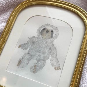 May include: Watercolor painting of a gray sloth wearing a hooded outfit, framed in a gold ornate frame. The sloth has a light gray body, a darker gray face, and brown feet. The name "Sammy" is written above the sloth.