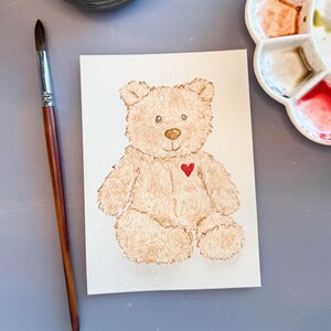 May include: Watercolor painting of a teddy bear with a red heart on its chest. The bear is light brown with a fluffy texture, sitting on a white paper. A paintbrush and a paint palette are in the background.
