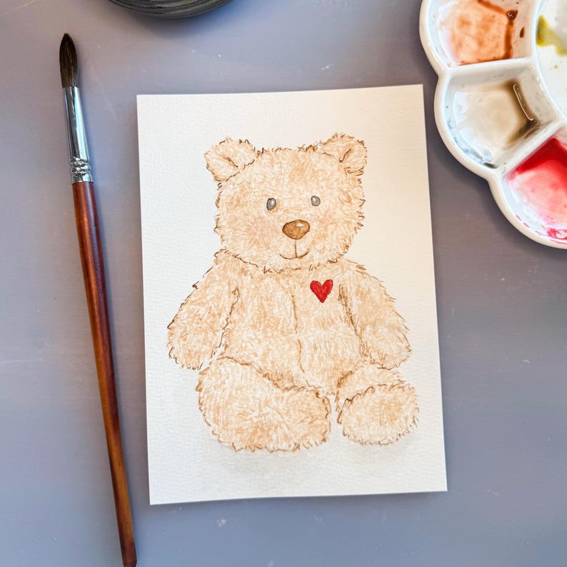 May include: Watercolor painting of a teddy bear with a red heart on its chest. The bear is light brown with a fluffy texture, sitting on a white paper. A paintbrush and a paint palette are in the background.