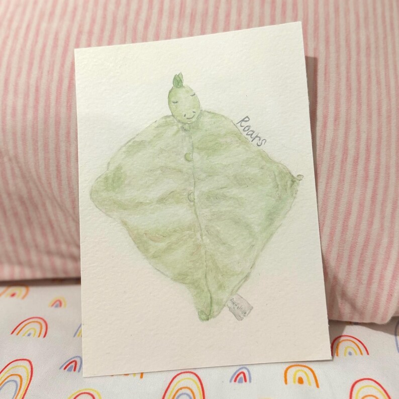 May include: Watercolor illustration of a green dinosaur-shaped baby blanket with button closures. The word "Roars" is written in cursive. The blanket is on a white card, set against a pink and white striped background.