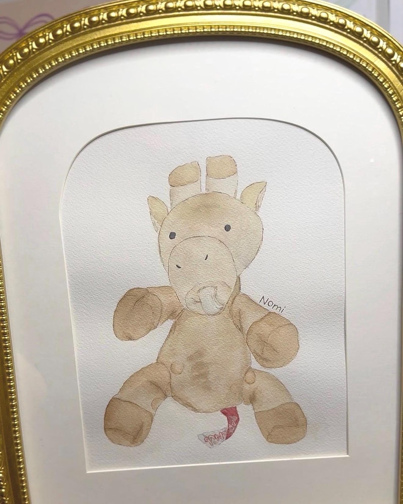 May include: Watercolor painting of a tan giraffe plush toy with a pacifier, framed in a gold ornate frame. The giraffe has brown accents and is sitting. The name "Nomi" is written on the right side of the painting.