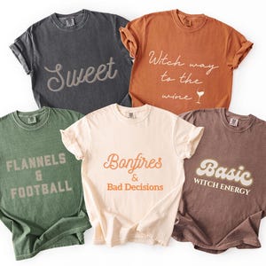 May include: A collection of five short-sleeved t-shirts in various colors, each with a unique graphic design. The shirts feature phrases like "Sweet," "Witch way to the wine," and "Bonfires & Bad Decisions."