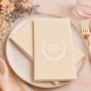 May include: A beige paper napkin folded on a speckled ceramic plate, featuring a white laurel wreath graphic surrounding the text BRING YOUR OWN LOGO. The napkin has a diagonal embossed border pattern at the bottom edge, placed next to gold cutlery.