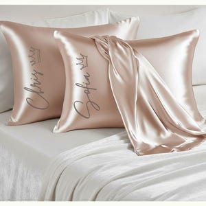 May include: Two blush-colored silk pillowcases with personalized names and crowns printed in a silver script. One pillow has the name "Chris" and the other "Sofia". A silk scarf is draped over the pillows on a white bed.