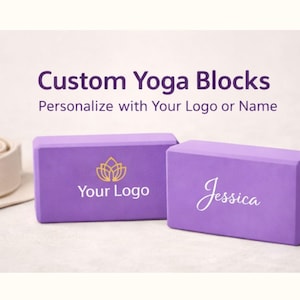 Op de afbeelding: Twee lavendelkleurige yogablokken met aangepaste tekst. Eén blok heeft een gouden lotusbloemlogo en de woorden "Your Logo". Het andere blok heeft de naam "Jessica" in witte letters. Een yogariem staat op de achtergrond. Bovenin staat de tekst "Custom Yoga Blocks".