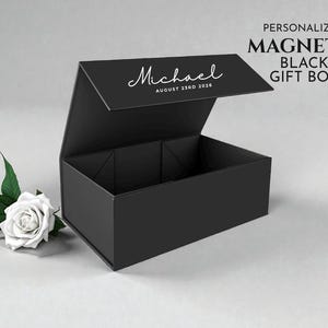 May include: A black magnetic gift box with the name "Michael" and date "August 23rd 2026" printed on the lid. A white rose and green leaves are next to the box. Text on the right says "PERSONALIZED MAGNETIC BLACK GIFT BOX".