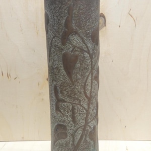 Puede incluir: Un jarrón cilíndrico de metal alto con un detallado patrón de hojas y vides. El jarrón tiene una apariencia oscura y envejecida con una superficie texturizada y un ligero tono azul-verdoso. El diseño está en relieve sobre el metal.