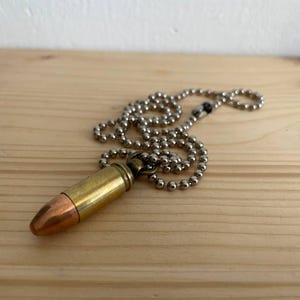 May include: A bullet pendant necklace featuring a brass and copper bullet on a silver-coloured ball chain. The bullet has a copper tip and brass casing. The necklace is displayed on a light brown wooden surface.