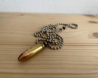 Könnte beinhalten: Halskette mit einem Anhänger in Form einer Kugel aus Messing und Kupfer an einer silberfarbenen Kugelkette. Die Kugel hat eine Kupferspitze und ein Messinggehäuse. Die Halskette liegt auf einer hellbraunen Holzoberfläche.
