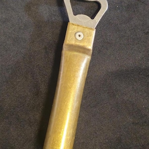 Puede incluir: Un abridor de botellas con un abridor plateado y un mango de latón con forma de casquillo de bala. El mango es cilíndrico y tiene un acabado metálico. El abridor está diseñado para abrir botellas.