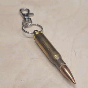 Op de afbeelding: Een messing kogel sleutelhanger met een zilverkleurige karabijnhaak en sleutelring. De kogel heeft een goudkleurige punt en een cilindrisch lichaam. De sleutelhanger is ontworpen om aan sleutels of een tas te worden bevestigd.