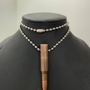 Pode incluir: Um colar com um pingente de bala cor de cobre suspenso de uma corrente de bolas prateada. O colar é exibido em um manequim preto. A bala tem uma ponta e um estojo metálico.