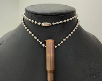 Könnte beinhalten: Eine Halskette mit einem kupferfarbenen Patronenanhänger, der an einer silbernen Kugelkette hängt. Die Halskette wird auf einer schwarzen Schaufensterpuppe präsentiert. Die Patrone hat eine Spitze und ein Metallgehäuse.