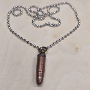 Op de afbeelding: Een koperkleurige kogelhangerketting. De kogelvormige hanger hangt aan een zilveren kogelketting. De hanger heeft een messingkleurige bovenkant en een zilverkleurige onderkant. De achtergrond is een lichte houtnerf.