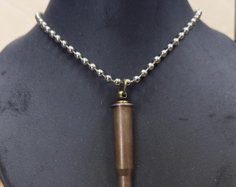 Può includere: Una collana con un ciondolo a proiettile. Il ciondolo è color rame con una fascia rossa vicino alla punta, sospeso a una catena a sfere argentata. La collana è esposta su un manichino nero.