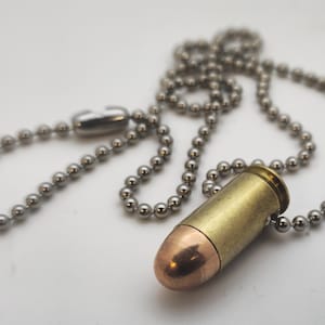 Puede incluir: Un collar con colgante en forma de bala con una carcasa de latón y punta de cobre. El colgante está unido a una cadena de bolas plateada. El collar se muestra sobre una superficie blanca.