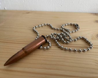 Könnte beinhalten: Eine kupferfarbene Halskette mit einem Anhänger in Form einer Kugel und einer silberfarbenen Kugelkette. Der kugelartige Anhänger liegt auf einer Holzoberfläche. Ein einzigartiges Accessoire.