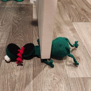 May include: A green and black crocheted doorstop shaped like a frog. The frog is lying on its side with its legs outstretched. The doorstop is placed against a white door.