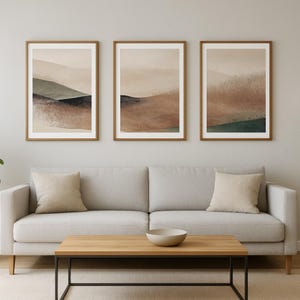 May include: Three framed abstract landscape art prints. Each print features a muted colour palette of brown, beige, and green. The artwork is displayed above a light grey sofa with beige cushions, a wooden coffee table, and a floor lamp.