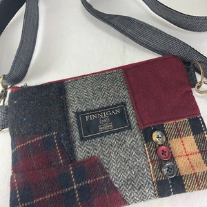 May include: A small, patchwork crossbody bag with a gray and red plaid pattern. The bag has a black strap and a label that reads "FINNIGAN Hand Crafted".
