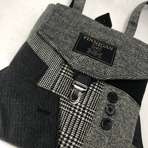 May include: A black and gray tweed backpack with a flap closure and a silver buckle. The backpack has a black label with the text "FINNIGAN Hand Crafted".