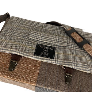 May include: A tweed messenger bag with a patchwork design. The bag features a mix of brown, gray, and tan fabrics, including a plaid pattern and a herringbone weave. It has a flap closure with a leather strap and a shoulder strap. The bag has a label that reads "FINNIGAN".
