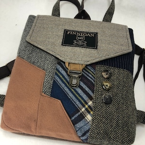 May include: A brown, gray, and blue patchwork backpack with a flap closure and a brown leather strap. The backpack has a label that reads "FINNIGAN" and "Hand Crafted".