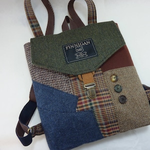 Pode incluir: Uma mochila patchwork com vários tecidos de tweed em tons de castanho, azul e verde. A aba tem uma correia e fivela de couro. A etiqueta diz "FINNIGAN".