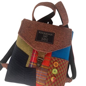May include: A brown, blue, green, and red patchwork backpack with a brown leather strap and buckle. The backpack has a label that says "Finnegan" and "Hand Crafted".