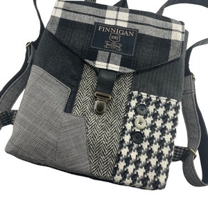 May include: Black and white plaid patchwork backpack with a leather flap and buckle closure. The bag has a small label that reads "Finnegan Sews Hand Crafted".