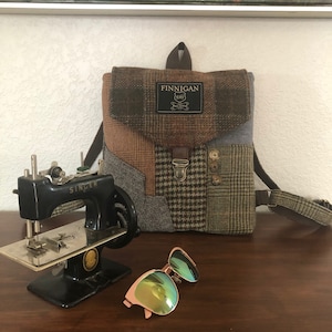 May include: A small, black Singer sewing machine sits next to a brown and green patchwork backpack with a leather strap and a label that reads "Finnegan 1845". A pair of pink sunglasses with green lenses are also in the image.