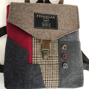 May include: A brown, gray, and red patchwork backpack with a brown leather strap and buckle. The backpack has a flap with a black label that reads "FINNIGAN" and "Hand Crafted".