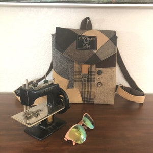 May include: A small brown and tan patchwork backpack with a brown leather strap and a label that reads "Finnegan 1865". The backpack is sitting on a wooden table next to a small black sewing machine and a pair of green sunglasses.