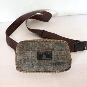 May include: A brown tweed fanny pack with a brown adjustable strap and a black buckle. The bag has a brown and tan trim and a label that reads "FINNIGAN".