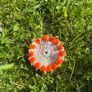 May include: A circular crochet coaster with a light beige centre and a border of bright orange pom-poms. The coaster is set on green grass, creating a natural and vibrant setting.