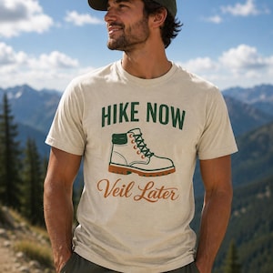 Peut inclure: T-shirt beige avec l'inscription "Hike Now Veil Later" en vert et orange. Le t-shirt présente un dessin d'une chaussure de randonnée. La personne sur l'image porte une casquette verte et se tient devant une chaîne de montagnes.