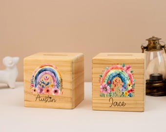 May include: Two wooden cube piggy banks. Each bank features a colorful rainbow design with floral accents. One bank has the name "Austin" and the other has "Jace" written on it. The banks are made of light-colored wood.
