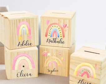May include: Wooden money boxes with rainbow designs in pink, yellow, and blue. Each box has a name printed on it: Addie, Nathalie, Olivia, Sophie, and Alicia. The boxes are made of light-colored wood.