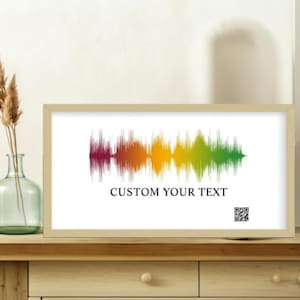 May include: A framed print featuring a colorful sound wave design. The sound wave transitions from burgundy to green, with text that reads "CUSTOM YOUR TEXT". The frame is a light beige color, and the print is displayed on a wooden surface.