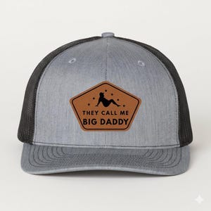 May include: Gray trucker hat with a black mesh back and a curved brim. The hat features a brown leather patch with the text "THEY CALL ME BIG DADDY" and a silhouette graphic.