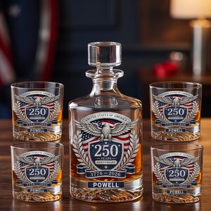 May include: A clear glass decanter with a stopper and four matching whiskey glasses, each featuring a United States of America 250th anniversary emblem with an eagle, a flag, the text 250 YEARS ANNIVERSARY 1776-2026, and the name POWELL.