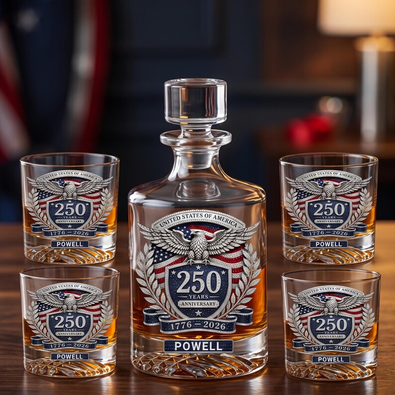 May include: A clear glass decanter with a stopper and four matching whiskey glasses, each featuring a United States of America 250th anniversary emblem with an eagle, a flag, the text 250 YEARS ANNIVERSARY 1776-2026, and the name POWELL.