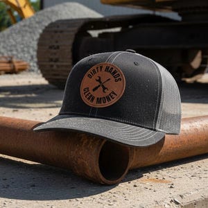 May include: A black and grey trucker hat featuring a circular brown leather patch on the front. The patch displays the text "DIRTY HANDS CLEAN MONEY" in black lettering, surrounding a graphic of two crossed wrenches in the center of the circle.