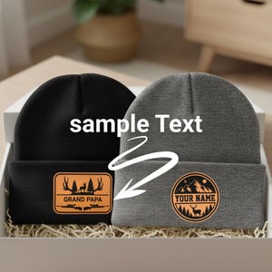 May include: A black beanie with a rectangular tan patch featuring a deer, trees, rifles, and the text GRAND PAPA, and a gray beanie with a circular tan patch featuring mountains, trees, a deer, and the text YOUR NAME, with sample Text overlay.